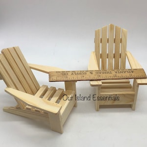 Puede incluir: Dos sillas Adirondack de madera en miniatura, una con una peque&ntilde;a mesa adjunta. Las sillas son de madera sin terminar y tienen aproximadamente 28 cm de altura. Las sillas est&aacute;n sobre una superficie blanca. El texto "Out Island Essentials" est&aacute; impreso en la superficie.
