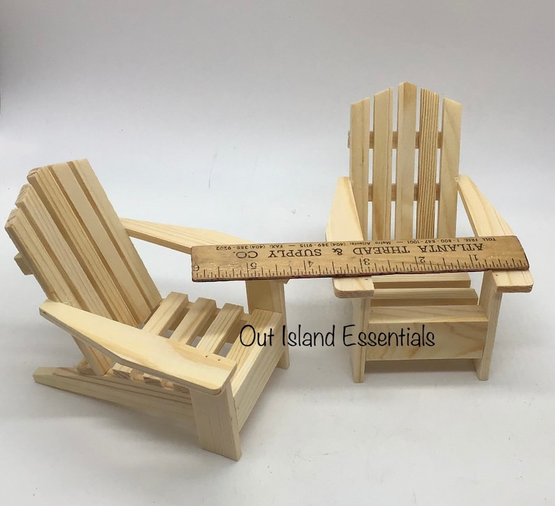 Puede incluir: Dos sillas Adirondack de madera en miniatura, una con una peque&ntilde;a mesa adjunta. Las sillas son de madera sin terminar y tienen aproximadamente 28 cm de altura. Las sillas est&aacute;n sobre una superficie blanca. El texto "Out Island Essentials" est&aacute; impreso en la superficie.