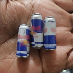 May include: Three small Red Bull energy drink cans. The cans are silver with blue and red branding. The text "ENERGY DRINK" is visible on one can. Each can is approximately 3 inches tall.