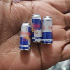 May include: Three small aluminum cans of energy drink, each with a blue and silver design and the words "ENERGY DRINK" visible. The cans are held in a hand.