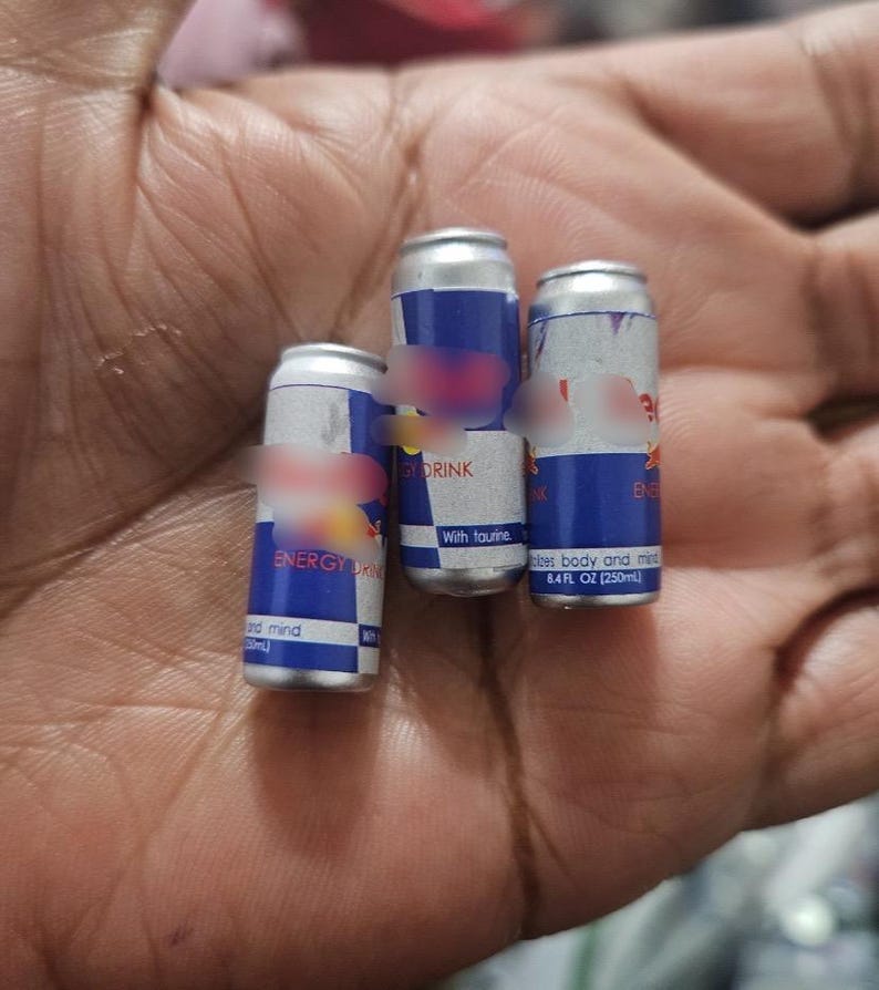 May include: Three small aluminum cans of energy drink, each with a blue and silver design and the words "ENERGY DRINK" visible. The cans are held in a hand.