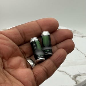 May include: Two miniature energy drink cans with a black and green design. The cans have a silver top and are held in a hand. The word "ENERGY" is visible on the can.