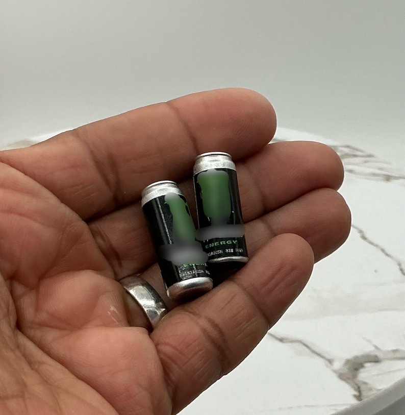 May include: Two miniature energy drink cans with a black and green design. The cans have a silver top and are held in a hand. The word "ENERGY" is visible on the can.