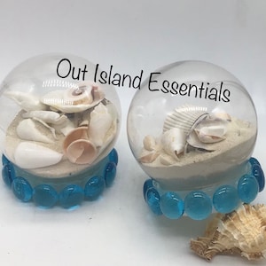 May include: Two clear glass globes filled with sand and seashells, each globe sits on a base of blue glass beads. The text "Out Island Essentials" is visible in the image.
