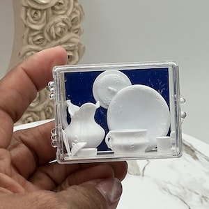 May include: A miniature white porcelain tea set, including a pitcher, plates, and cups, is displayed inside a clear plastic case. The set has a delicate floral design. The background is a dark blue color.