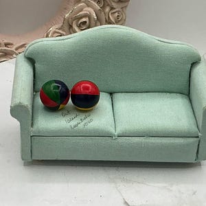 May include: A light green miniature sofa with two small, colorful balls on the seat. The sofa has a curved back and a handwritten message on the seat that reads "Out Island Essentials xoxo".