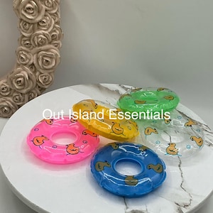 Könnte beinhalten: Eine Sammlung bunter, aufblasbarer Schwimmreifen in Pink, Gelb, Grün, Blau und Transparent. Jeder Reifen hat ein sich wiederholendes Muster von Cartoon-Enten. Die Reifen sind auf einer weißen Oberfläche mit Marmormuster angeordnet. Der Text "Out Island Essentials" ist sichtbar.