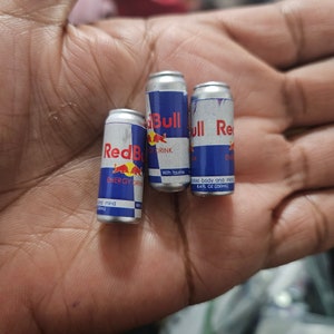 May include: Three miniature Red Bull energy drink cans. The cans are silver with blue and red branding, featuring the Red Bull logo and text. Each can is labeled with "ENERGY DRINK" and "8.4 FL OZ (250ml)".
