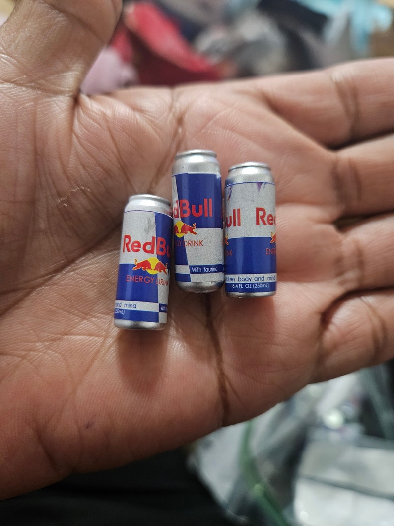 May include: Three miniature Red Bull energy drink cans. The cans are silver with blue and red branding, featuring the Red Bull logo and text. Each can is labeled with "ENERGY DRINK" and "8.4 FL OZ (250ml)".