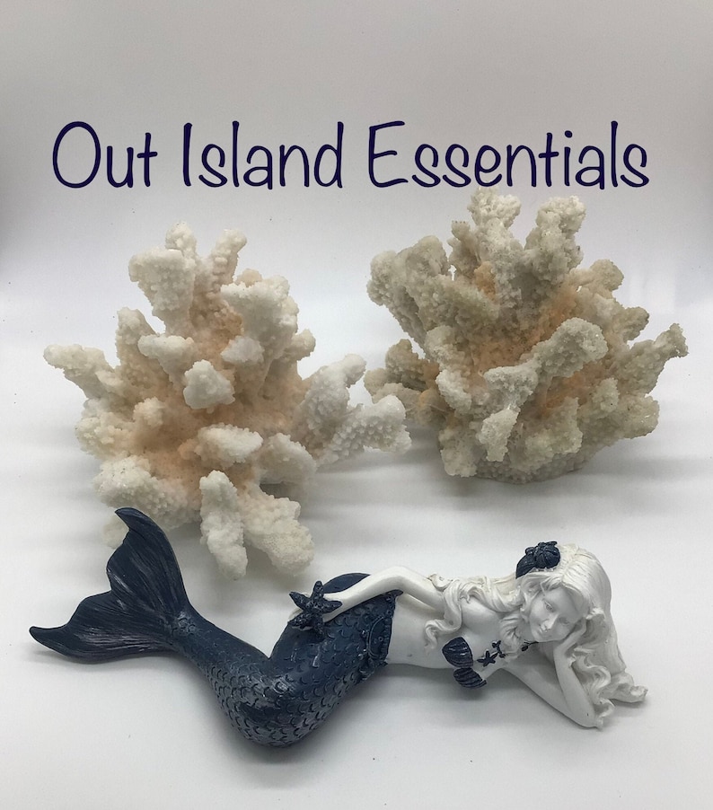 Pu&ograve; includere: Due decorazioni di barriera corallina bianca e una statuetta di sirena bianca con coda blu, sdraiata su una superficie bianca. Il testo "Out Island Essentials" &egrave; in alto nell'immagine.