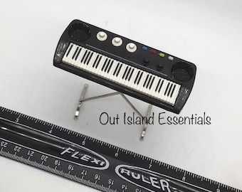 May include: A miniature black keyboard with white keys and silver stand. The keyboard has white knobs and speakers on each side. The text "Out Island Essentials" is visible in the image. A black ruler is in the foreground.