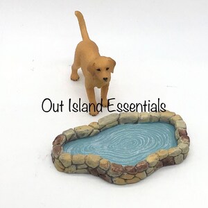 Puede incluir: Un conjunto de figuras en miniatura con un perro Labrador Retriever dorado y un pequeño estanque. El estanque está rodeado por un borde de piedra y lleno de agua azul. El texto "Out Island Essentials" está impreso en la imagen.