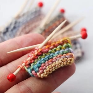May include: A miniature knitting project in progress, featuring a small swatch of colorful yarn in a rainbow pattern. The project is held with wooden knitting needles with red tips. The yarn colors include red, orange, yellow, green, blue, and purple.