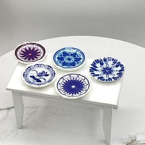 May include: A collection of miniature porcelain plates with blue and white floral patterns, displayed on a small white table. One plate is oval-shaped with a purple floral design. The other plates are round.
