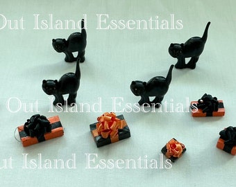Könnte beinhalten: Ein Set mit Miniatur-schwarzen Katzen und orange-schwarzen Geschenkboxen mit schwarzen Schleifen. Die Geschenkboxen sind mit orange-schwarzem Band verziert. Der Text "Out Island Essentials" erscheint dreimal im Bild.