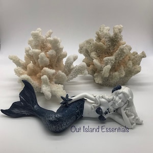Pu&ograve; includere: Una statuetta di sirena bianca con una coda blu, sdraiata su un fianco con la testa appoggiata sulla mano. Tiene una piccola stella marina nell'altra mano. Due pezzi di corallo bianco sono dietro di lei.