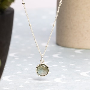 May include: A silver necklace with a round, iridescent labradorite gemstone pendant. The necklace has a delicate chain with small silver beads.