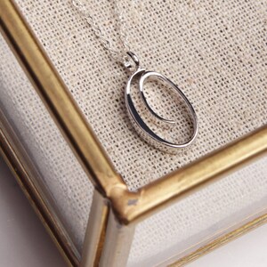 May include: A silver-colored pendant necklace featuring an open oval design with a curved inner detail. The necklace hangs on a delicate chain. The jewelry is displayed inside a clear glass box with a gold-colored frame, set against a neutral fabric background.