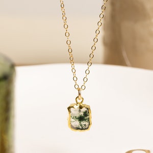 May include: A gold-colored necklace with a rectangular pendant. The pendant features a moss agate stone with green and white patterns, encased in a gold-colored bezel. The necklace is displayed on a white plate.