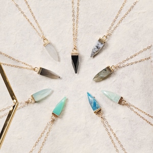 May include: A collection of ten gold necklaces with different colored gemstone pendants. The pendants are all shaped like arrowheads and are arranged in a circle on a white background.