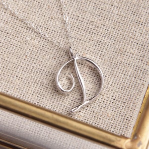 May include: A silver necklace with a cursive letter D pendant. The necklace is displayed on a beige fabric background.