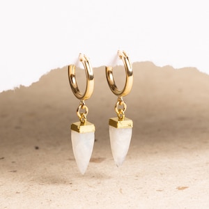 May include: Gold hoop earrings with a white gemstone pendant in the shape of a point. The earrings are on a white background.