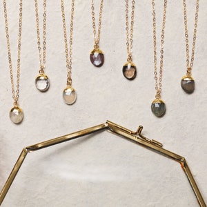 May include: Seven gold necklaces with different colored gemstone pendants. The pendants are all faceted and have a geometric shape. The necklaces are displayed on a white surface.