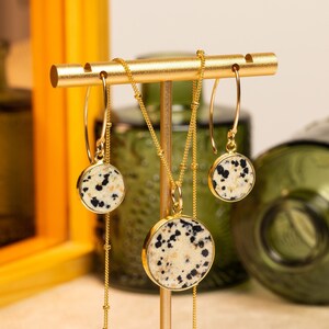 K&ouml;nnte beinhalten: Ein goldenes Halsketten- und Ohrring-Set mit einem Anh&auml;nger aus Dalmatiner-Jaspis und passenden Ohrringen. Die Halskette und die Ohrringe werden auf einem goldenen St&auml;nder pr&auml;sentiert.