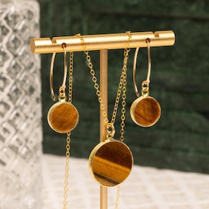 May include: A gold necklace and earrings set with round tiger's eye stone pendants. The necklace and earrings are displayed on a gold stand.