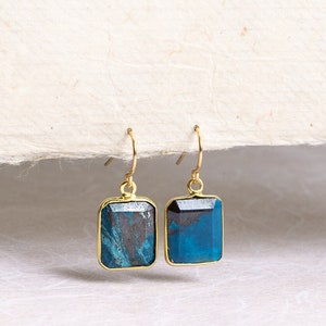 May include: A pair of gold earrings with blue and black gemstone drops. The earrings have a rectangular shape and are suspended from a gold hook.