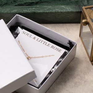 May include: A white box with a gold chain necklace inside. The box has a black velvet lining and a white card with the text "SOUL & LITTLE ROSE" printed on it.