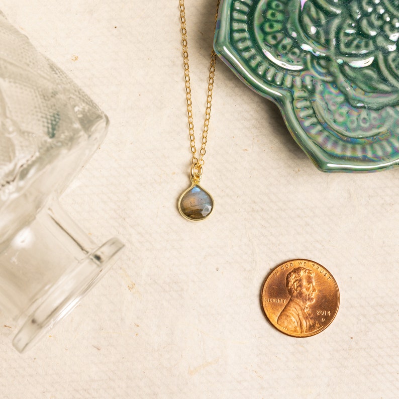 May include: A gold chain necklace with a teardrop-shaped labradorite gemstone pendant. The pendant is a light blue, gray, and brown color.