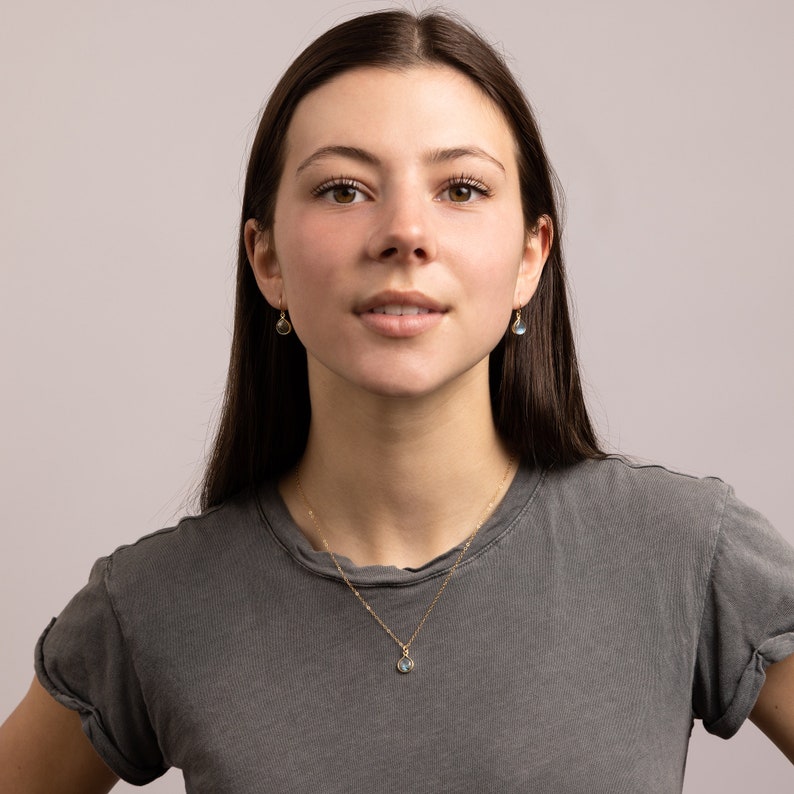 May include: A woman with long brown hair wearing a gray t-shirt and a gold necklace with a small blue gemstone pendant. She is also wearing small gold earrings with blue gemstones.
