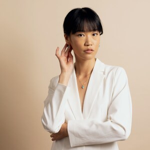 Puede incluir: Una mujer con cabello oscuro lleva una chaqueta blanca y un collar de oro con un colgante turquesa. Se est&aacute; tocando la oreja con la mano.