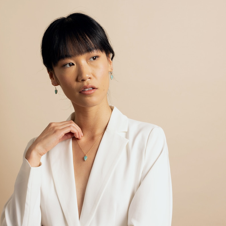 Puede incluir: Una mujer con cabello oscuro lleva una chaqueta blanca y un collar de oro con un colgante de piedra azul claro. Lleva peque&ntilde;os pendientes azules claros.