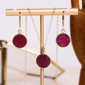 May include: A gold necklace and earrings set with a round, dark pink pendant and matching earrings. The jewelry is displayed on a gold stand.