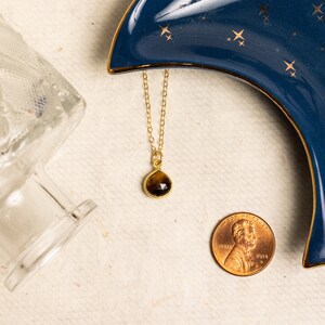 May include: A gold chain necklace with a teardrop-shaped brown gemstone pendant. The necklace is displayed on a blue and gold crescent moon-shaped dish with gold stars.