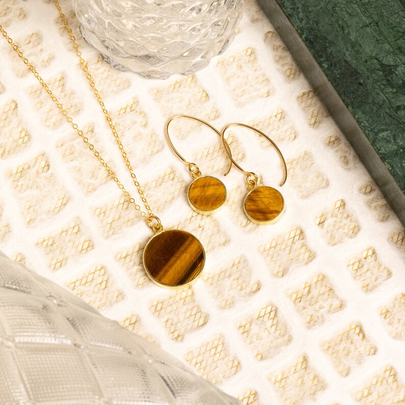 May include: A gold necklace and earrings set with tiger's eye stone pendants. The round pendants have a brown and gold color.