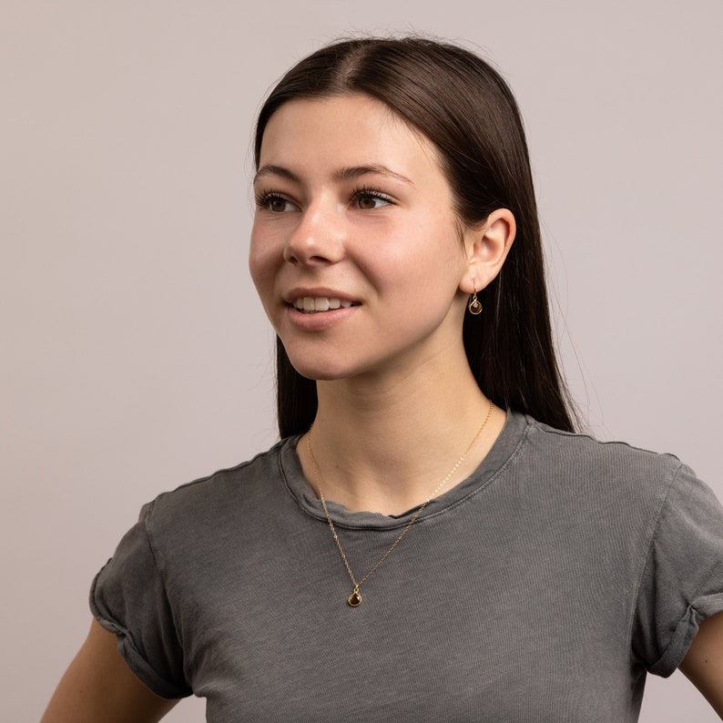 May include: A woman with long brown hair wearing a gray t-shirt and a gold necklace with a small teardrop pendant. She is also wearing gold earrings with teardrop stones.