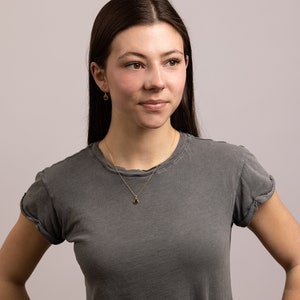May include: A woman wearing a gray t-shirt with a gold chain necklace and a small gold pendant. She is wearing small gold earrings.
