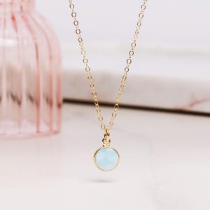 May include: A delicate gold chain necklace with a light blue gemstone pendant. The pendant is round and has a smooth, polished finish.