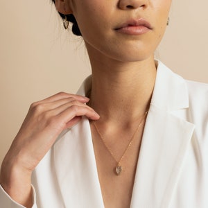May include: A gold chain necklace with a small, clear crystal pendant shaped like a leaf. The necklace is worn by a person wearing a white blazer.