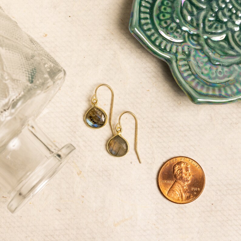 May include: A pair of gold dangle earrings with teardrop-shaped labradorite stones. The earrings are on a white surface with a green decorative dish and a US penny in the background.