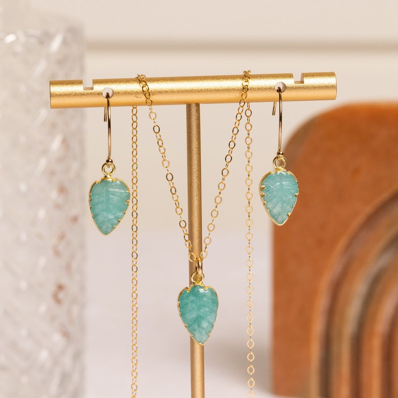 Puede incluir: Un collar y pendientes de oro con colgantes en forma de hoja de color verde turquesa. Las joyas se muestran en un soporte dorado.