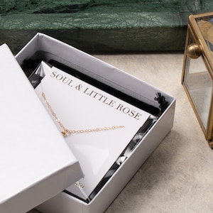 May include: A white box with a black velvet interior. The box contains a gold chain necklace with a small pendant. The box is labeled "Soul & Little Rose."