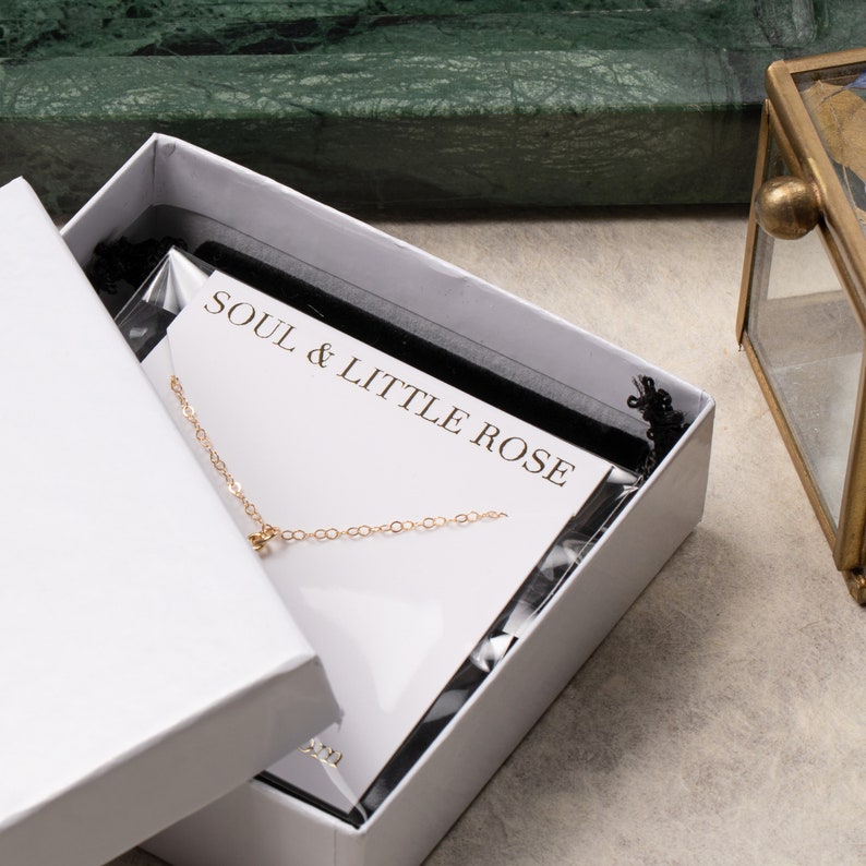 May include: A white box with a black velvet interior. The box contains a gold chain necklace with a small pendant. The box is labeled "Soul & Little Rose."