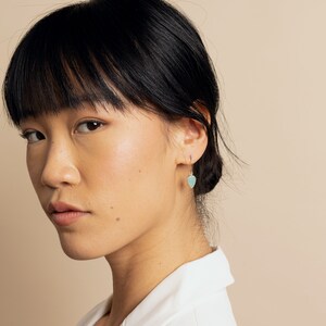 Puede incluir: Una mujer con cabello oscuro lleva un par de aretes de oro con una piedra de color azul claro en forma de l&aacute;grima.