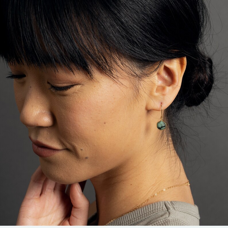 May include: A woman wearing a green gemstone earring with a gold setting. The earring is a simple drop style with a hexagonal stone.