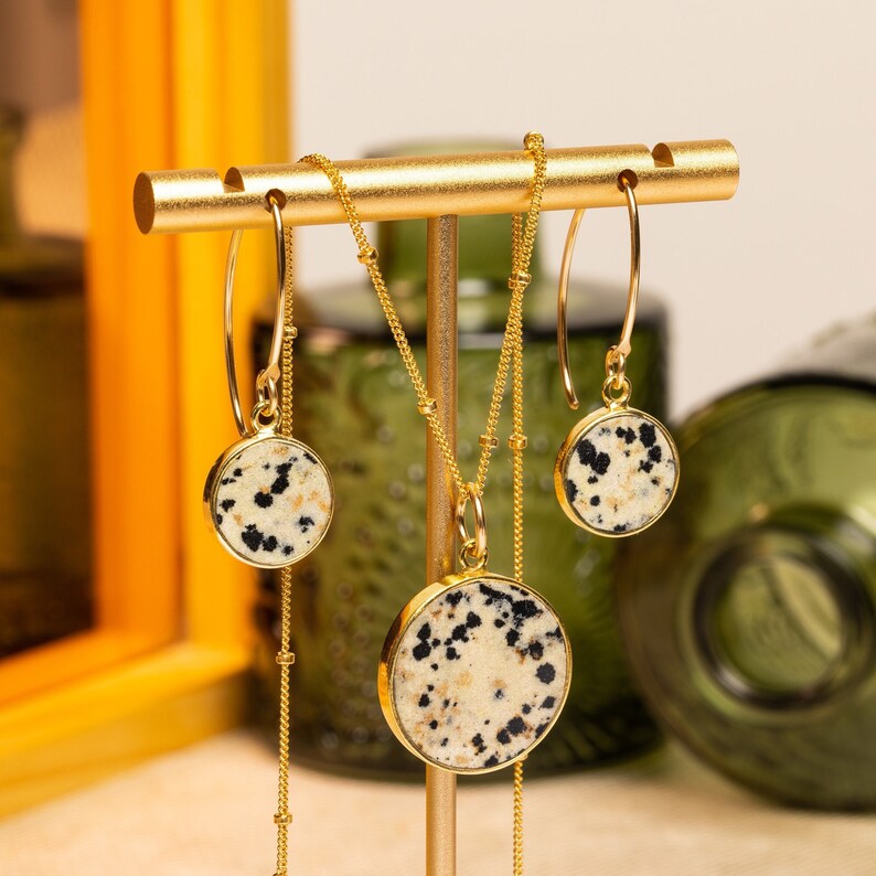 K&ouml;nnte beinhalten: Ein goldenes Halsketten- und Ohrring-Set mit einem Anh&auml;nger aus Dalmatiner-Jaspis und passenden Ohrringen. Die Halskette und die Ohrringe werden auf einem goldenen St&auml;nder pr&auml;sentiert.