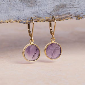 May include: A pair of gold-colored drop earrings. Each earring features a round, light purple gemstone set in a gold-tone bezel. The earrings have a lever-back closure. The background is a soft, neutral color.